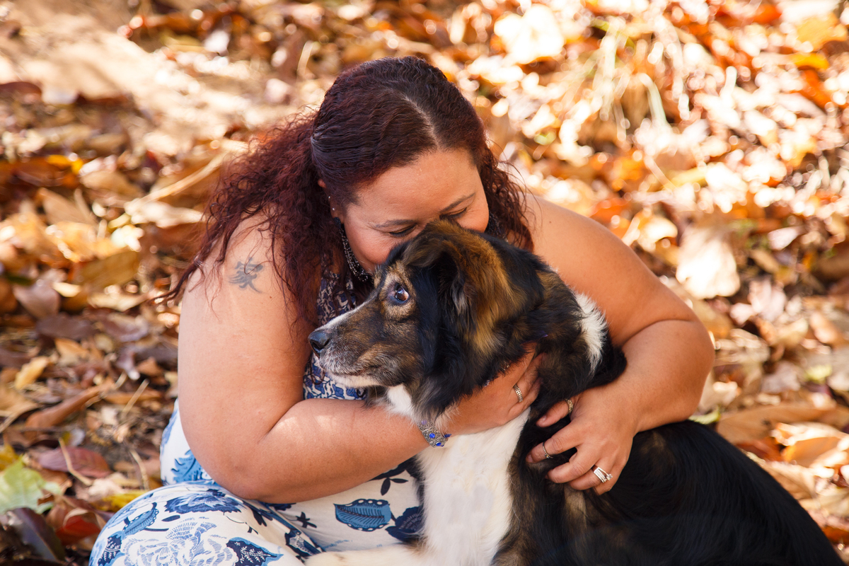 Mulher sentada no chão coberto de folhas secas abraçando seu cão border collie e dando um cheiro em sua cabeça.