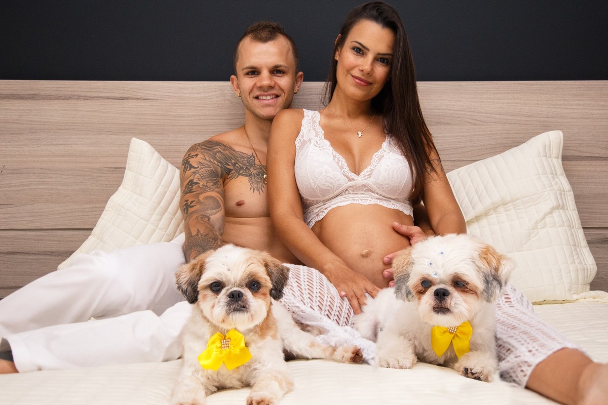 Família completa sentada na cama, mãe gestante, com o pai ao lado e as duas cachorrinhas da família, da raça shih tzu deitadas na cama à frente deles.