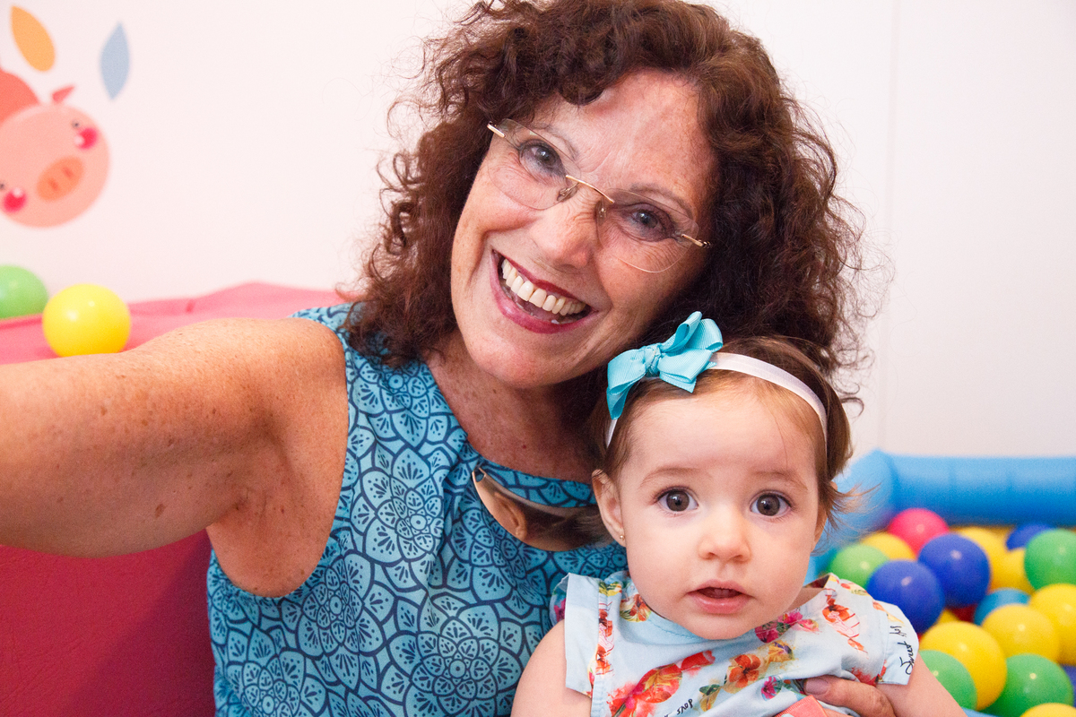 Avó posando para foto com sua netinha, em seu quarto com brinquedos ao fundo.