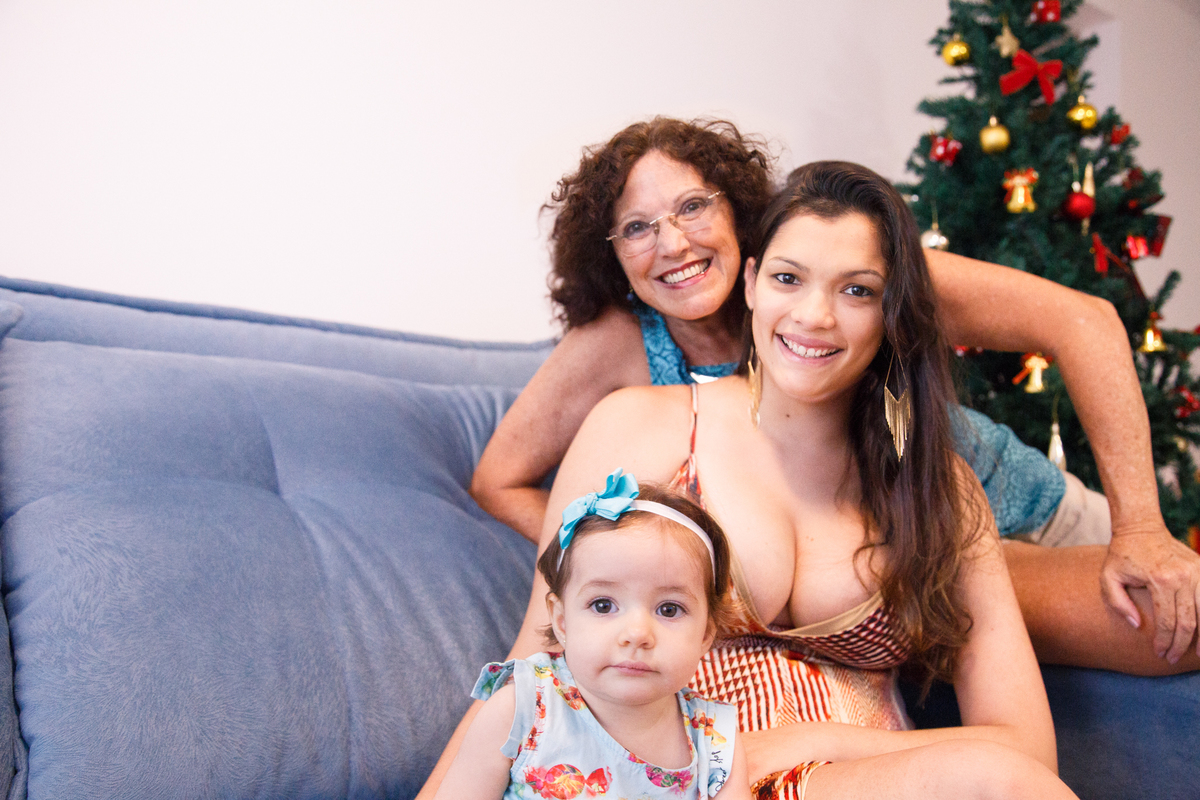 Três gerações de mulheres da família, avó, mãe e bebê, sentadas no sofá de casa e todas olhando para a câmera.