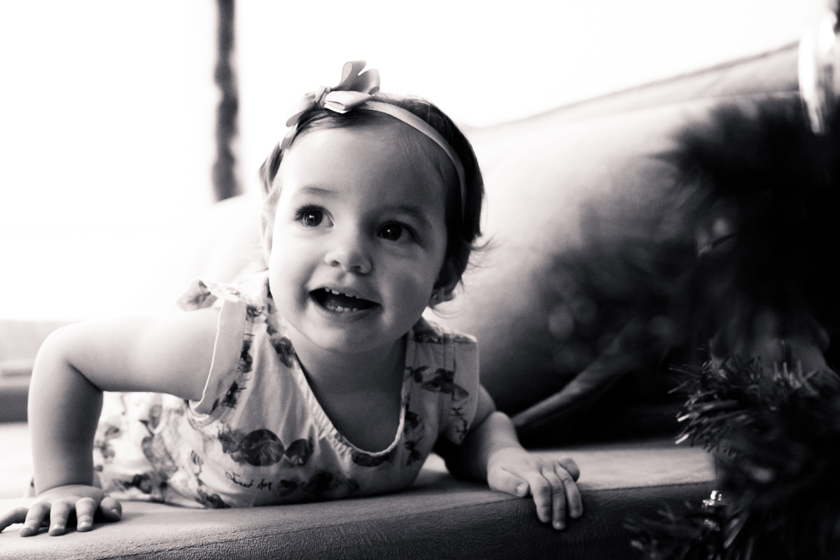 Foto em preto e branco de uma bebê debruçada no braço do sofá e sorrindo para a mãe, que não aparece na foto.