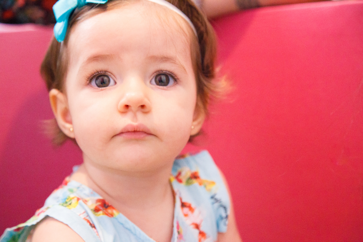 Close no rosto de uma bebê com lacinho azul, olhando para a foto, séria.