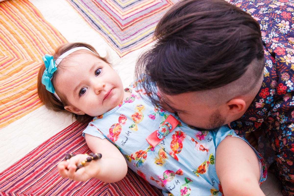 Bebê deitada na cama dos pais olhando para a câmera que está em um ângulo de cima, com o pai fazendo cócegas em sua barriga.
