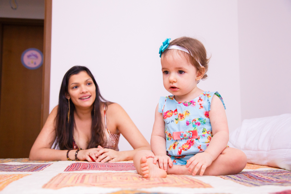 Em um primeiro plano uma bebê de vestidinho azul sentada na cama dos pais com a mãe ao fundo olhando para ela e sorrindo.