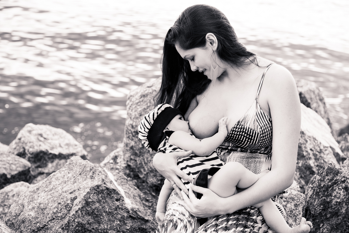 Foto em preto e branco de uma mãe amamentando sua bebê sentada nas pedras na beira da praia.