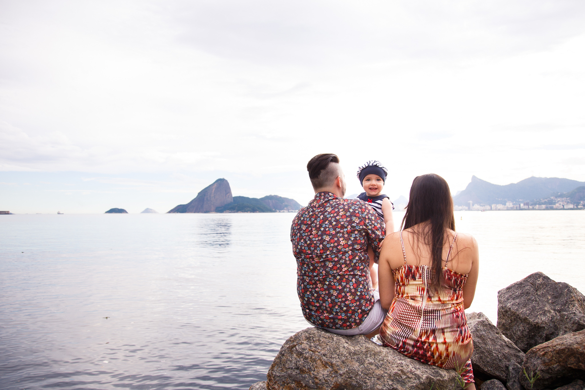 Mãe e pai sentados em pedras na beira da praia, de costas para a foto enquanto a bebê entre eles, no colo olha de frente para a câmera. Ao fundo a paisagem do Rio de Janeiro, com o Pão de Açúcar.