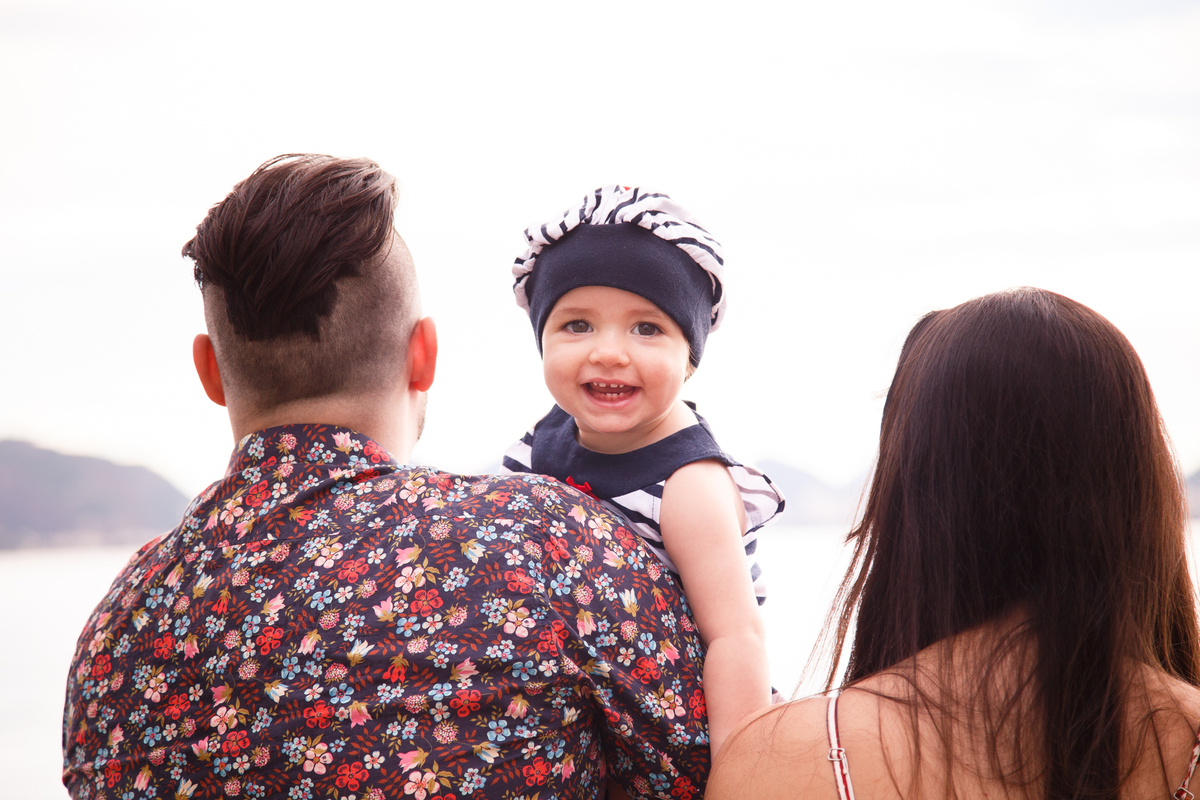 Close nas costas do pai e da mãe com a bebê entre eles olhando para a foto e sorrindo, usando um chapeuzinho listrado em azul e branco estilo marinheiro.