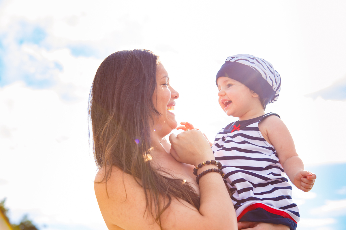 Bebê no colo de sua mãe, com o sol refletindo nelas, deixando um leve flare, o céu ao fundo e as duas sorrindo uma para a outra.