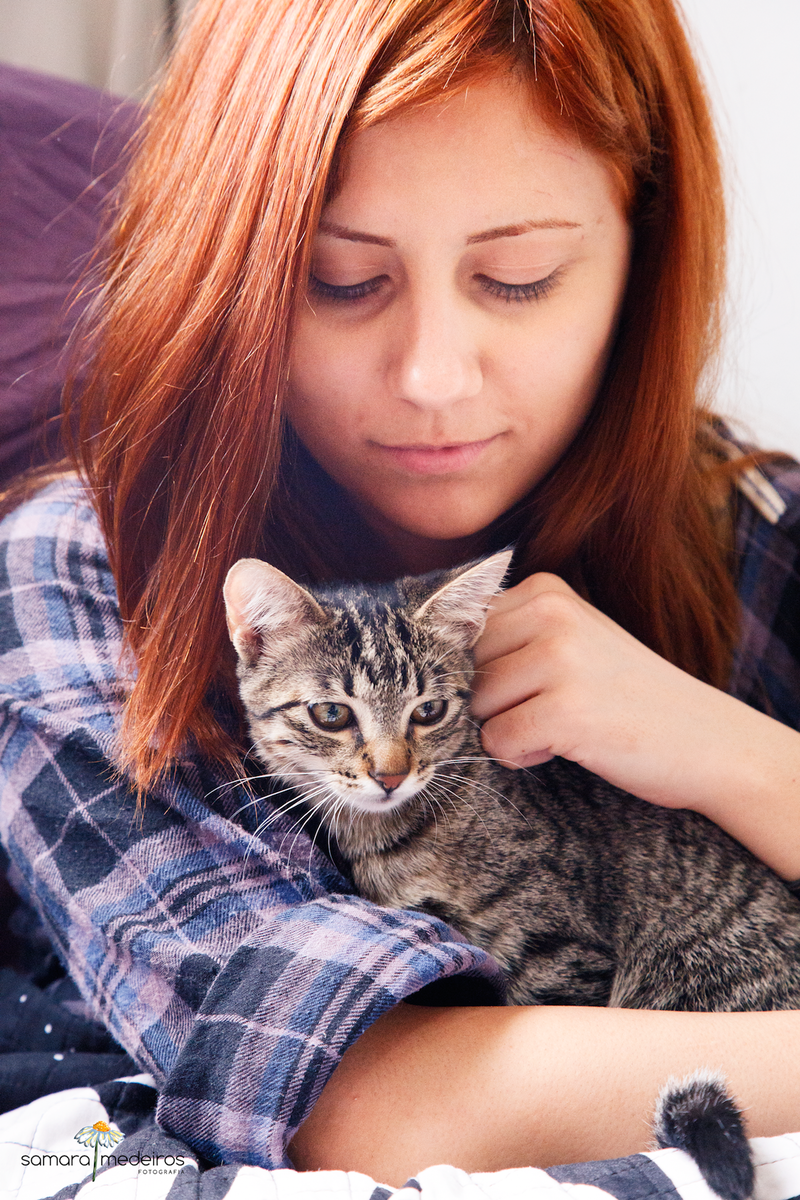 Close em meio rosto de uma moça ruiva e sua gatinha tigrada no colo.