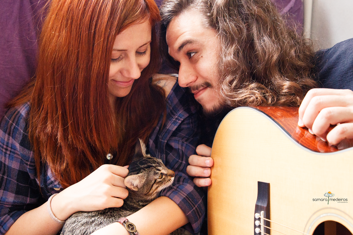 Casal sentado na cama com um violão deixado de lado, enquanto o rapaz olha a moça, a moça olha a gatinha tigrada em seu colo, todos sorrindo.