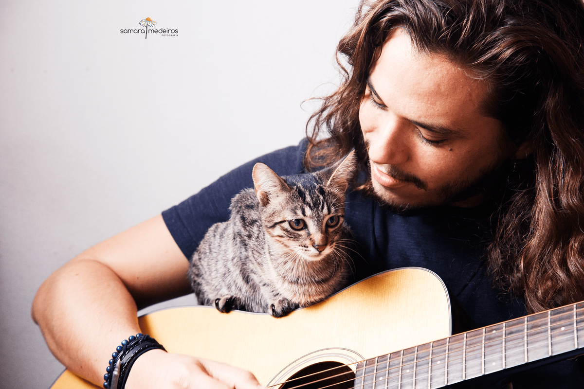 Close em um rapaz tocando violão com sua gatinha tigrada em cima do violão, enquanto ele olha para ela.