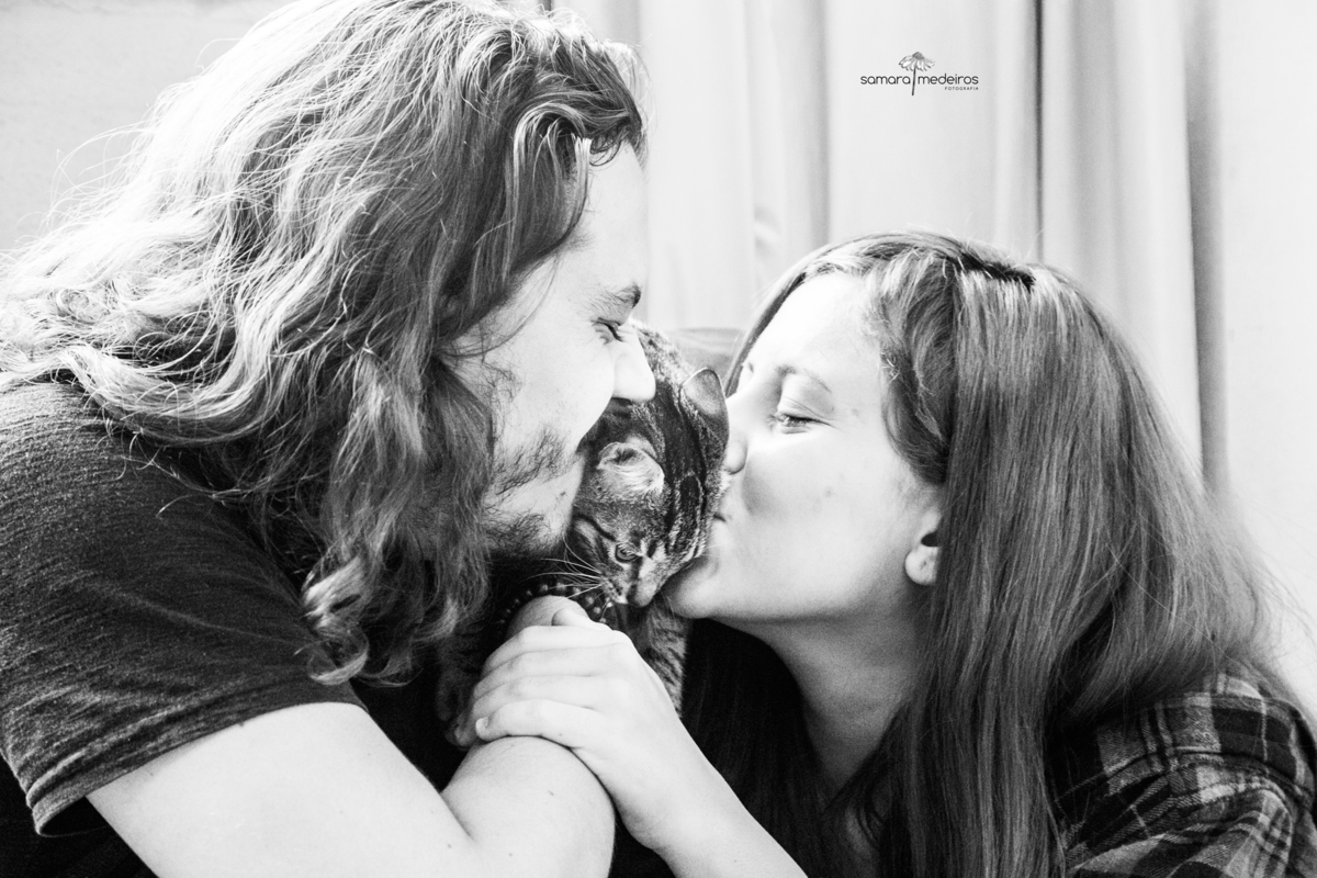 Foto em preto e branco de um casal beijando sua gatinha tigrada, um de cada lado dela.