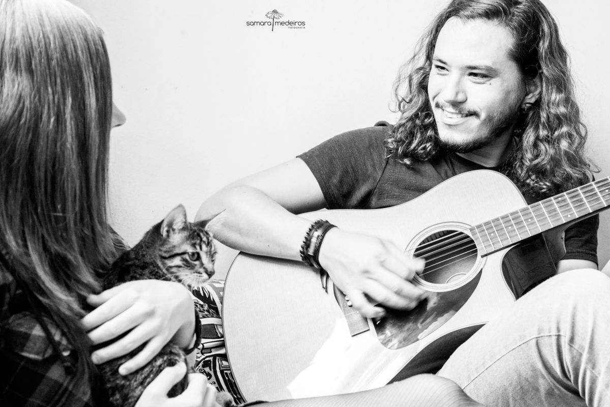 Casal sentado na cama, o rapaz tocando violão e olhando para a moça sorrindo, que o observa com uma gatinha tigrada no colo.