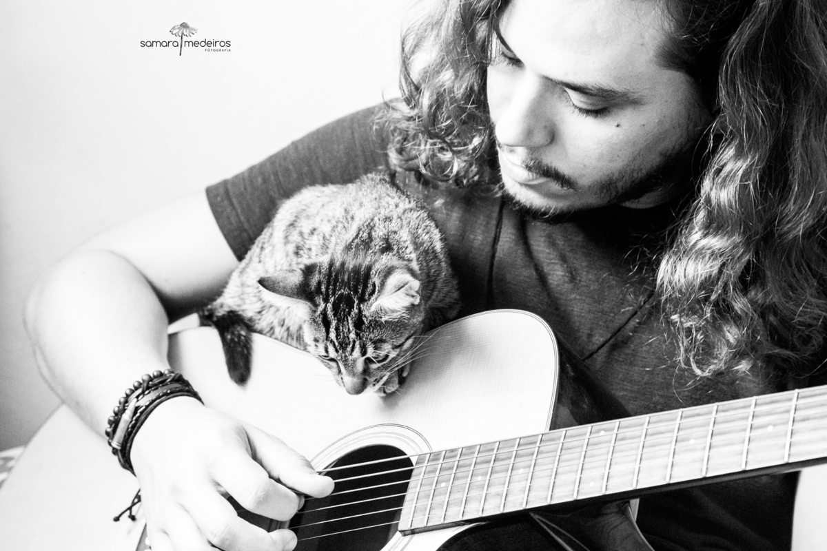 Foto em preto e branco de um rapaz tocando com uma gatinha tigrada em cima do violão. Ela olha para a mão dele e ele olha para ela.