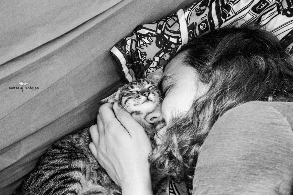 Foto em preto e branco de um rapaz deitado na cama abraçando sua gatinha tigrada, que faz uma expressão como se sorrisse.