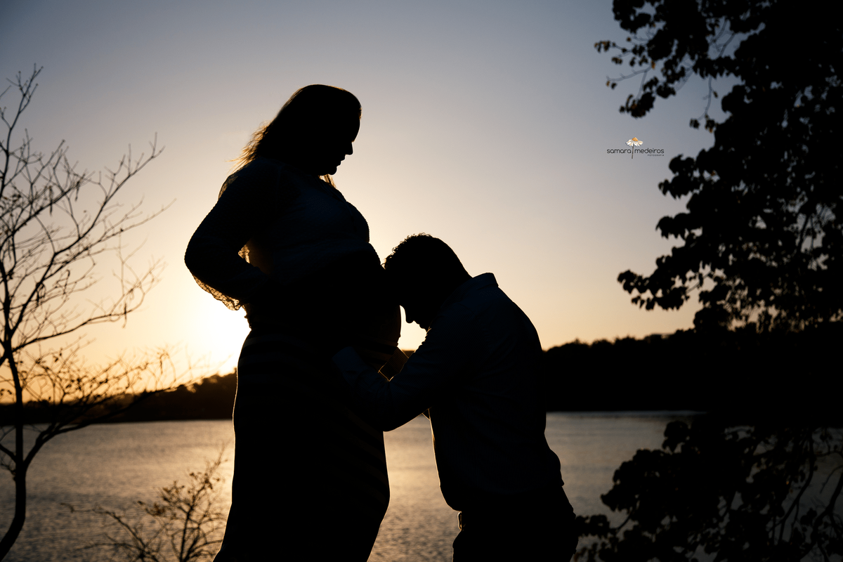 Gestante em silhueta com a lagoa da Pampulha ao fundo e o marido com a cabeça apoiada na barriga.