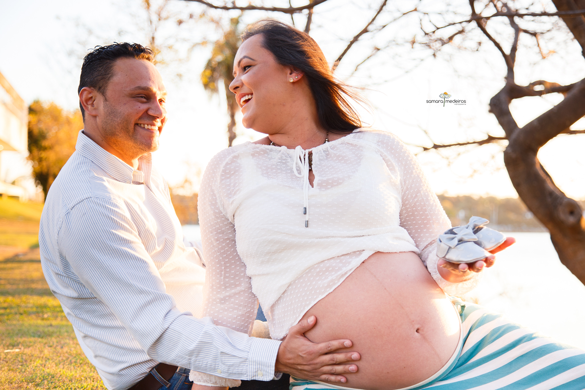 Casal esperando um bebê sentados na grama na beira da lagoa sorrindo um para o outro, a mãe com um par de sapatinhos na mão.