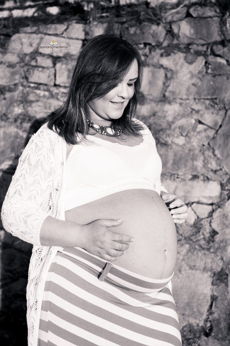 Foto em preto e branco de uma gestante encostada em uma parede de pedras, olhando e acariciando a barriga.