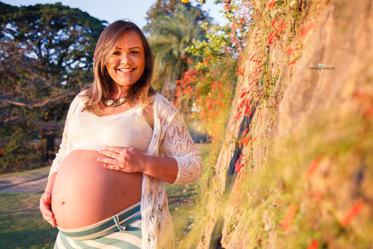 Gestante ao lado de uma parede de pedras com algumas flores, olhando para a câmera e sorrindo.