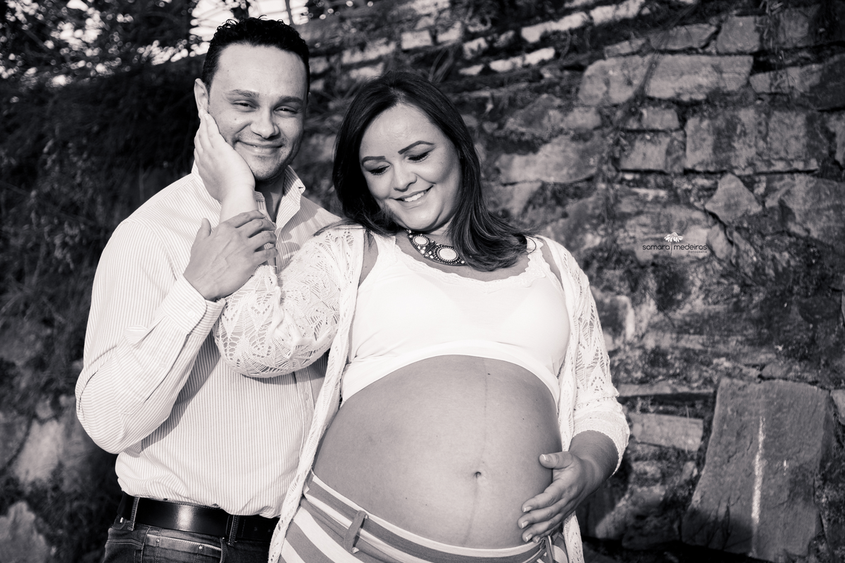 Foto em preto e branco de um casal esperando um bebê, o homem olhando para a câmera enquanto a mulher olha e acaricia a barriga e o rosto do marido.