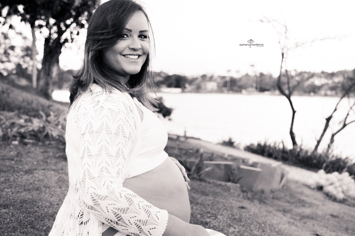Foto em preto e branco de uma gestante sentada na grama na orla da lagoa da pampulha, tendo a paisagem da orla ao fundo.