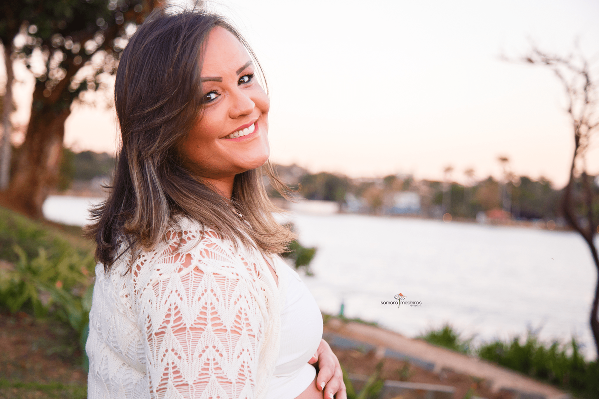 Gestante sentada na grama na orla da Lagoa da Pampulha acariciando a barriga e olhando para trás, para a foto.