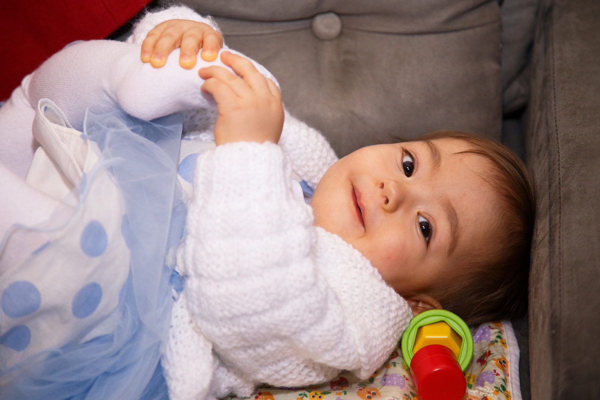 Bebê de 1 ano em seu aniversário infantil deitada no sofá mexendo no pé.