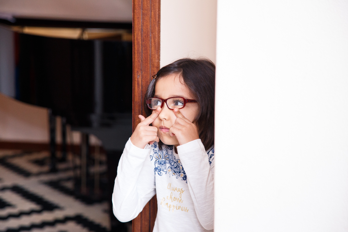 Criança apoiada em uma parede observando o ambiente e com as duas mãos empurrando as lentes do óculos.