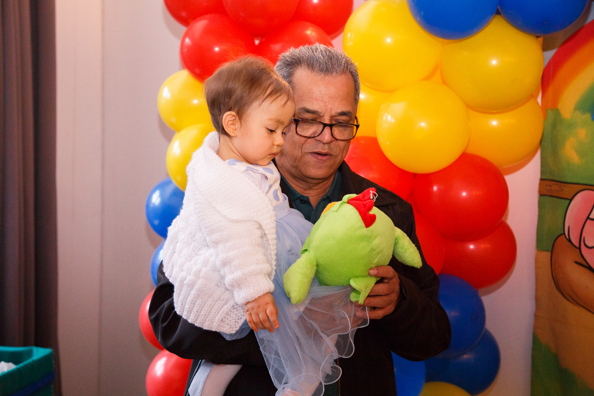 Bebê de 1 ano no colo do avó vendo uma galinha de pelúcia durante seu aniversário em Belo Horizonte.