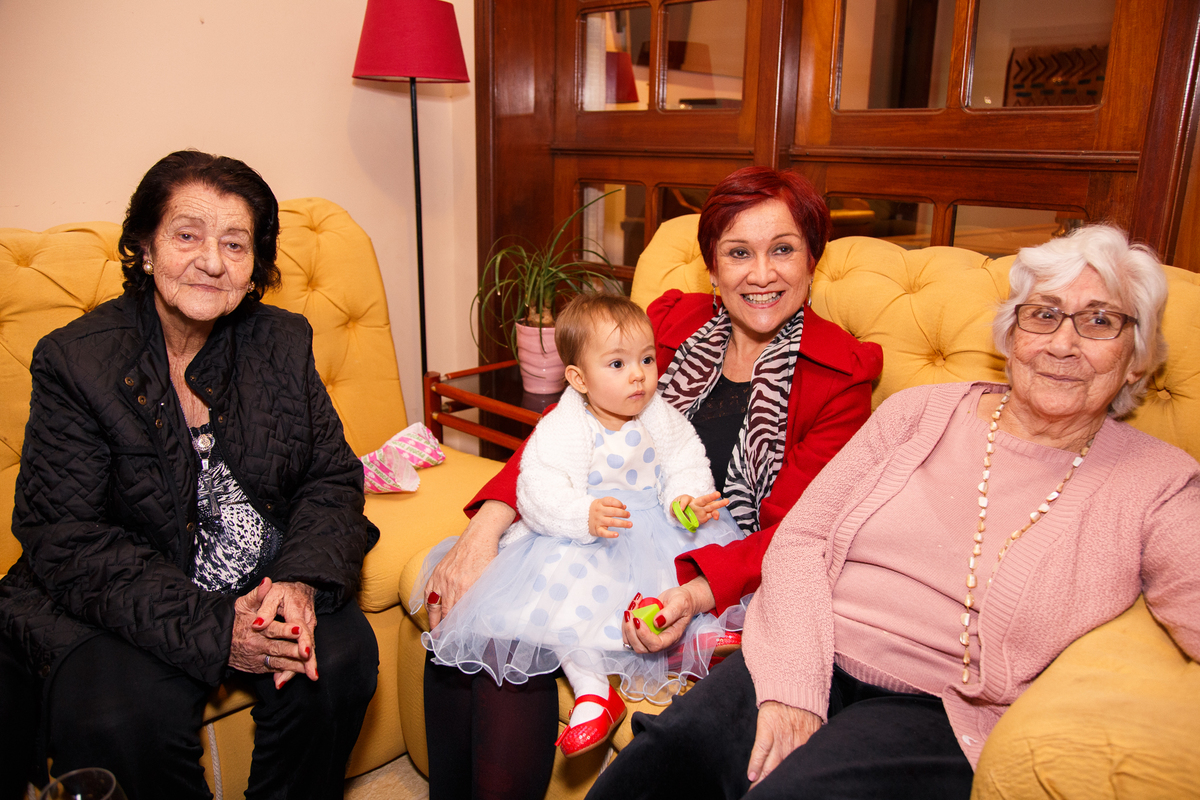 Bebê em seu aniversário de 1 ano posando para foto com sua avó e duas bisavós, sentadas em um sofá.