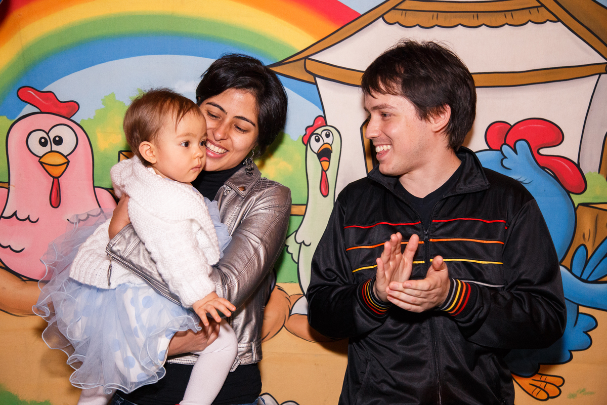 Pai, mãe e bebê atrás da mesa do bolo cantando parabéns durante aniversário infantil.