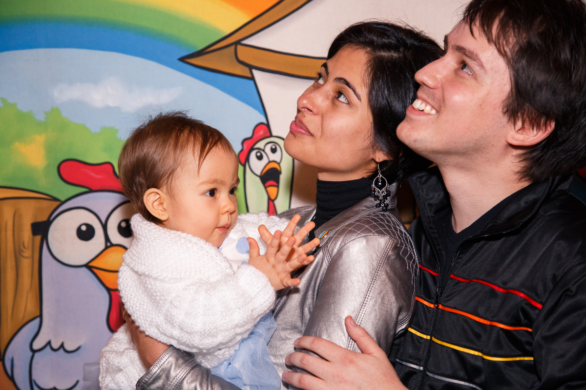 Bebê batendo palminhas no colo de sua mãe, com seu pai abraçando elas, e os dois olhando para cima para assistir um vídeo slideshow.