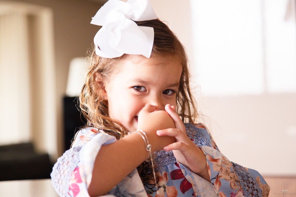 Close no rosto de uma criança que está rindo com a mão cobrindo a boca e olhando para a câmera.