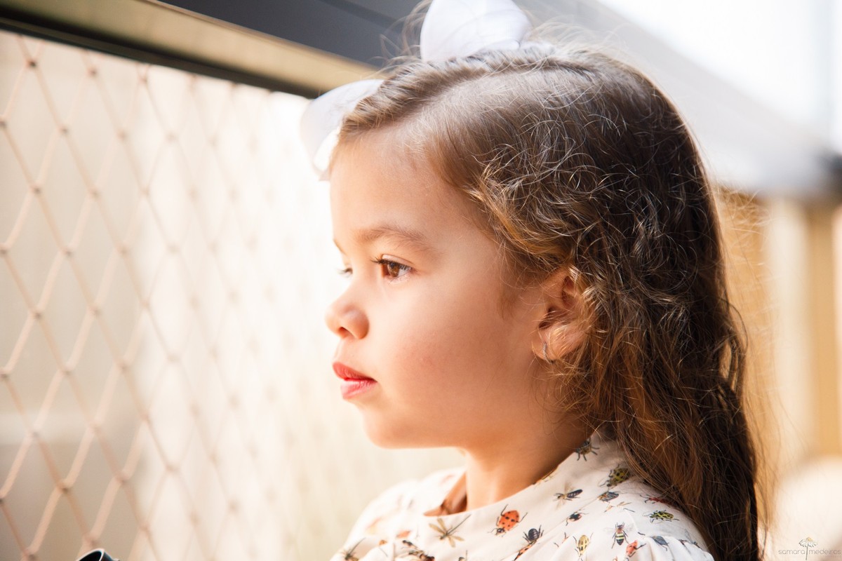 Criança séria olhando pela janela de sua casa durante sessão de foto infantil.