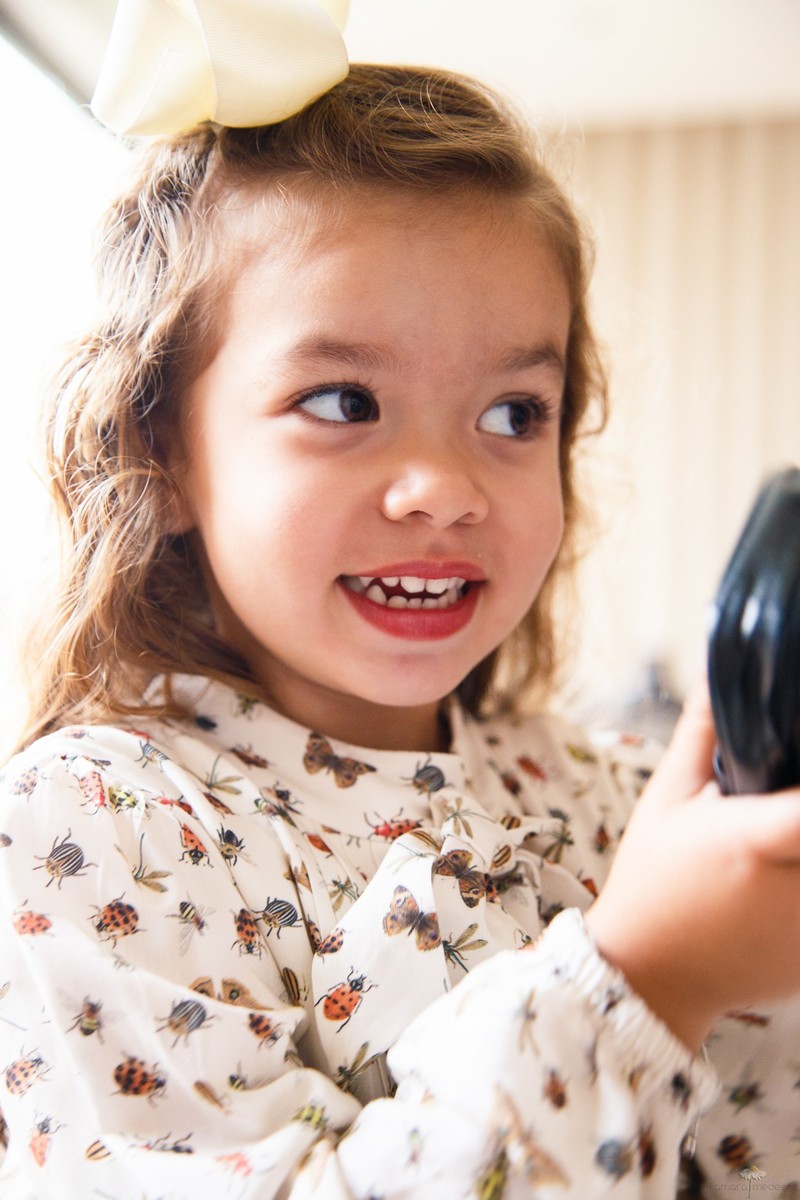 Close no rosto de uma criança sorrindo para a mãe, que não aparece na foto, segurando uma bolsinha de couro.