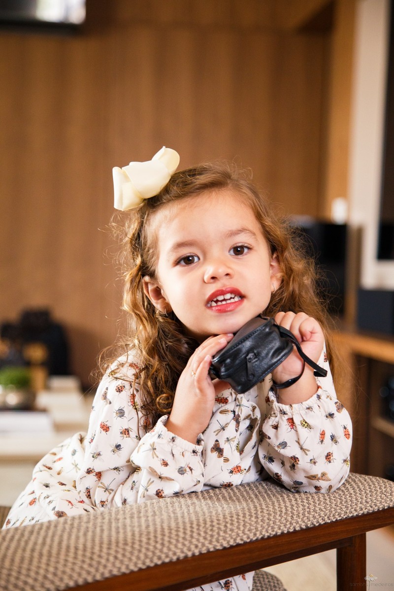 Criança segurando seu binóculo de brinquedo, dentro da capa de couro, debruçada no braço do sofá e olhando para a foto.