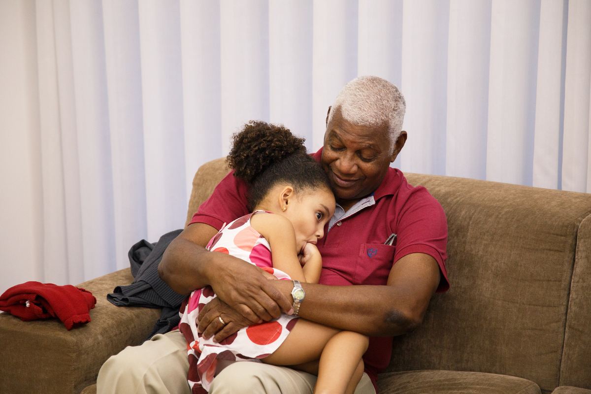 Criança sentada no sofá no colo do avô em aniversário infantil da irmã de 1 ano.