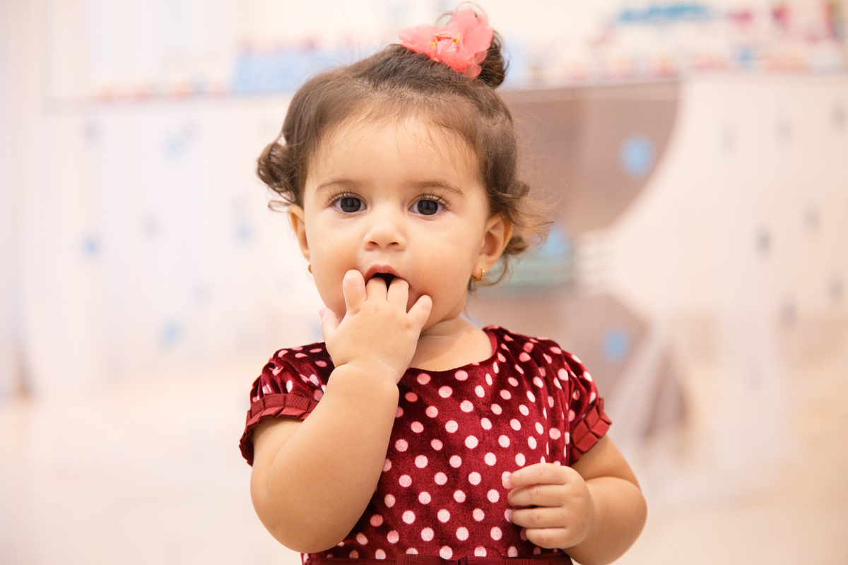 Bebê em frente à mesa do bolo de seu aniversário de 1 ano com a mão na boca, olhando para a câmera.