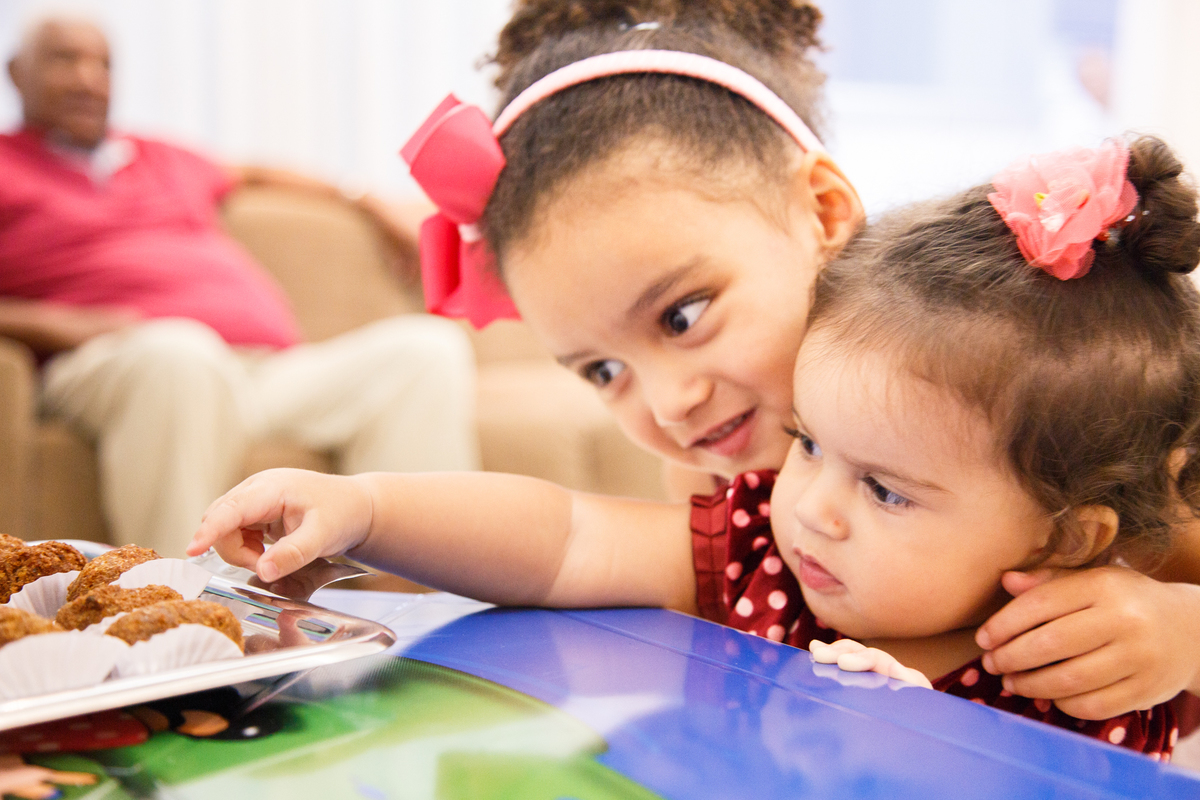 Bebê esticando o bracinho para pegar um salgado enquanto sua irmã mais velha está abraçando ela por trás e sorrindo.