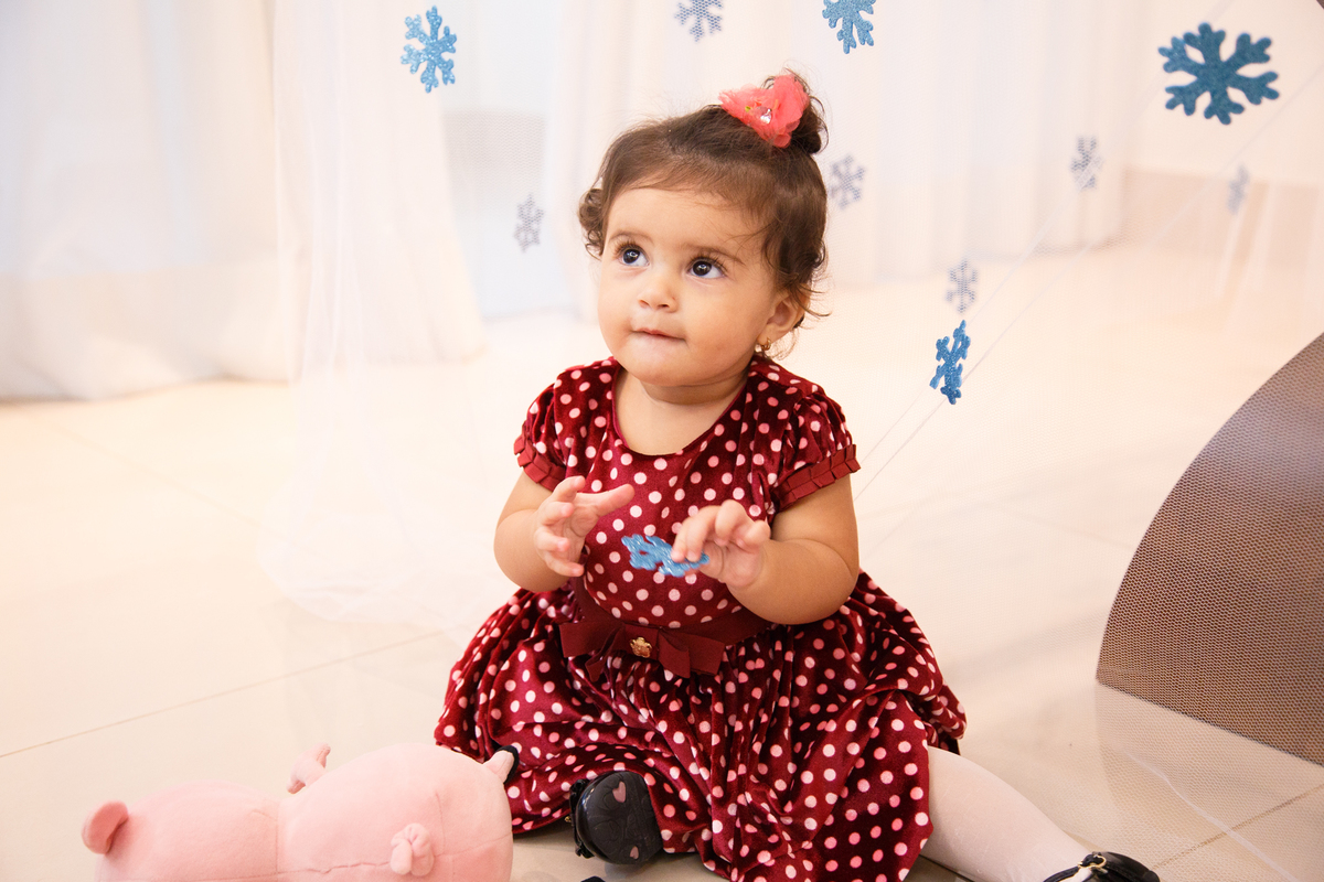 Bebê aniversariante em seu aniversário de 1 ano, sentada no chão, brincando com os flocos de neve de decoração no tema Frozen.