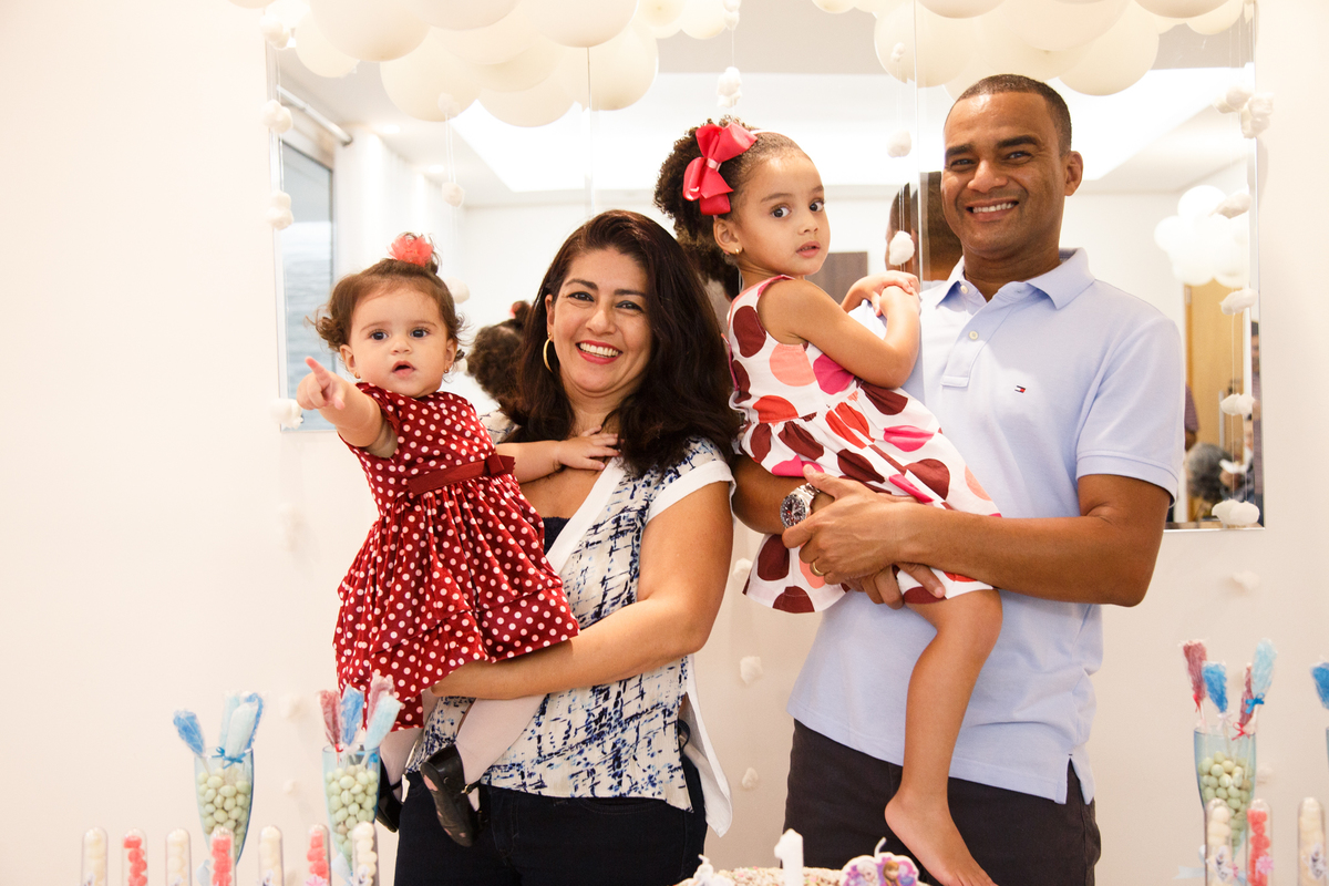 Pais com suas filhas no colo, atrás da mesa do bolo no aniversário de 1 ano da mais nova.