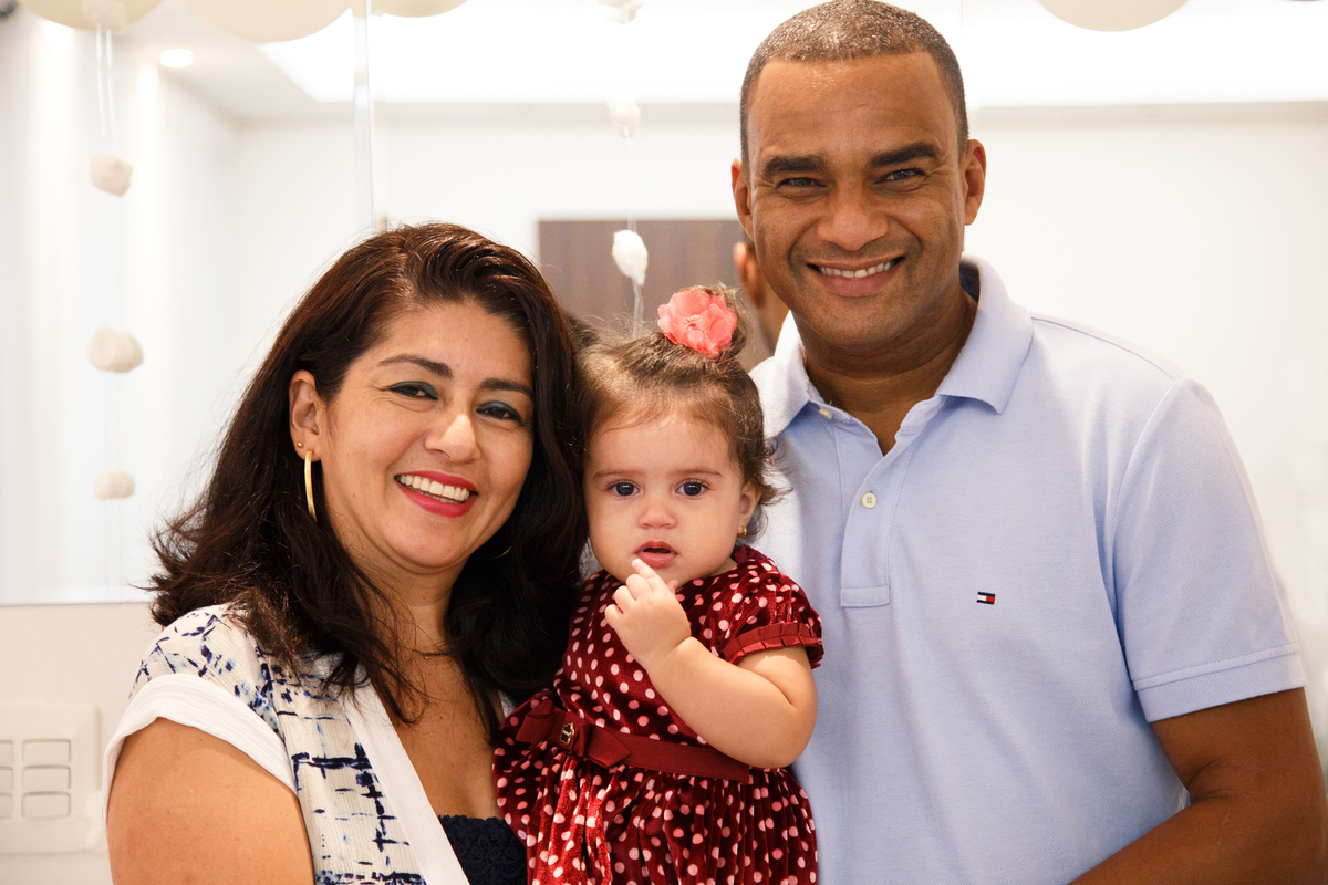 Pai e mãe com sua bebê no colo posando para foto no aniversário de 1 ano da criança.