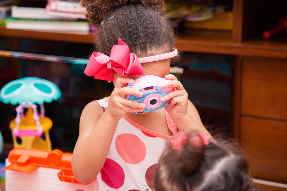 Criança brincando com uma câmera fotográfica de brinquedo, fazendo uma foto da sua irmã mais nova, que está fazendo aniversário de 1 ano.