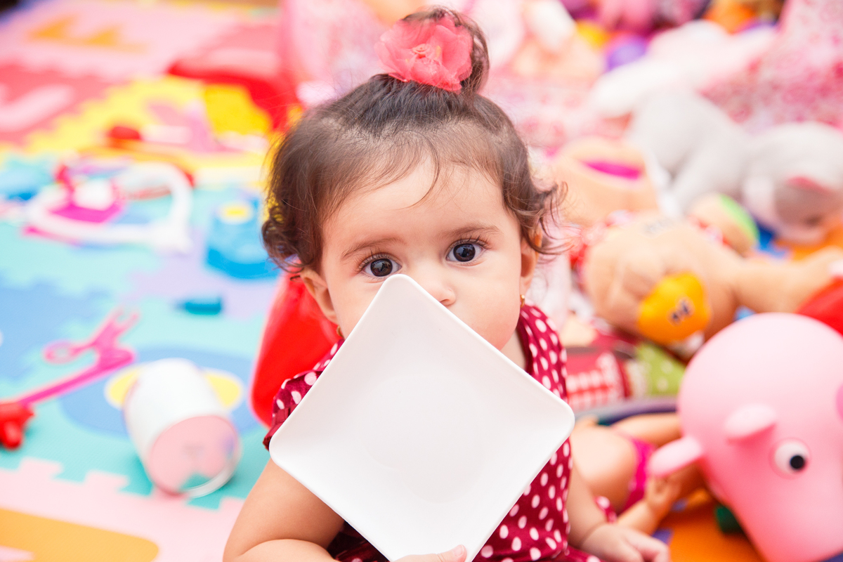 Bebê sentada no quarto dos brinquedos com um pratinho de plástico parcialmente em frente ao rosto, com o foco em seus olhos.