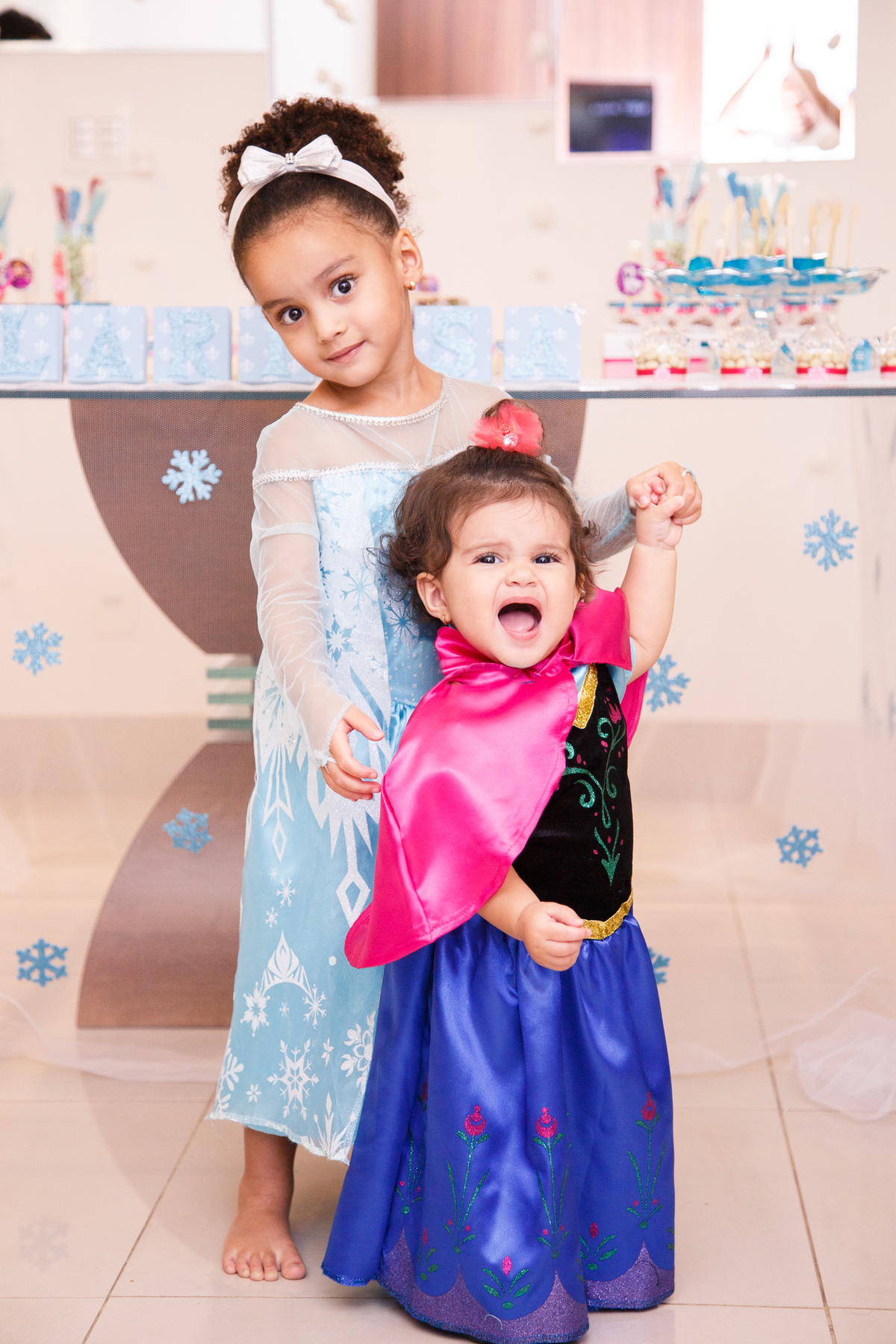 Duas irmãs fantasiadas de Anna e Elsa no aniversário de um ano da criança mais nova, as duas em frente à mesa do bolo sorrindo.