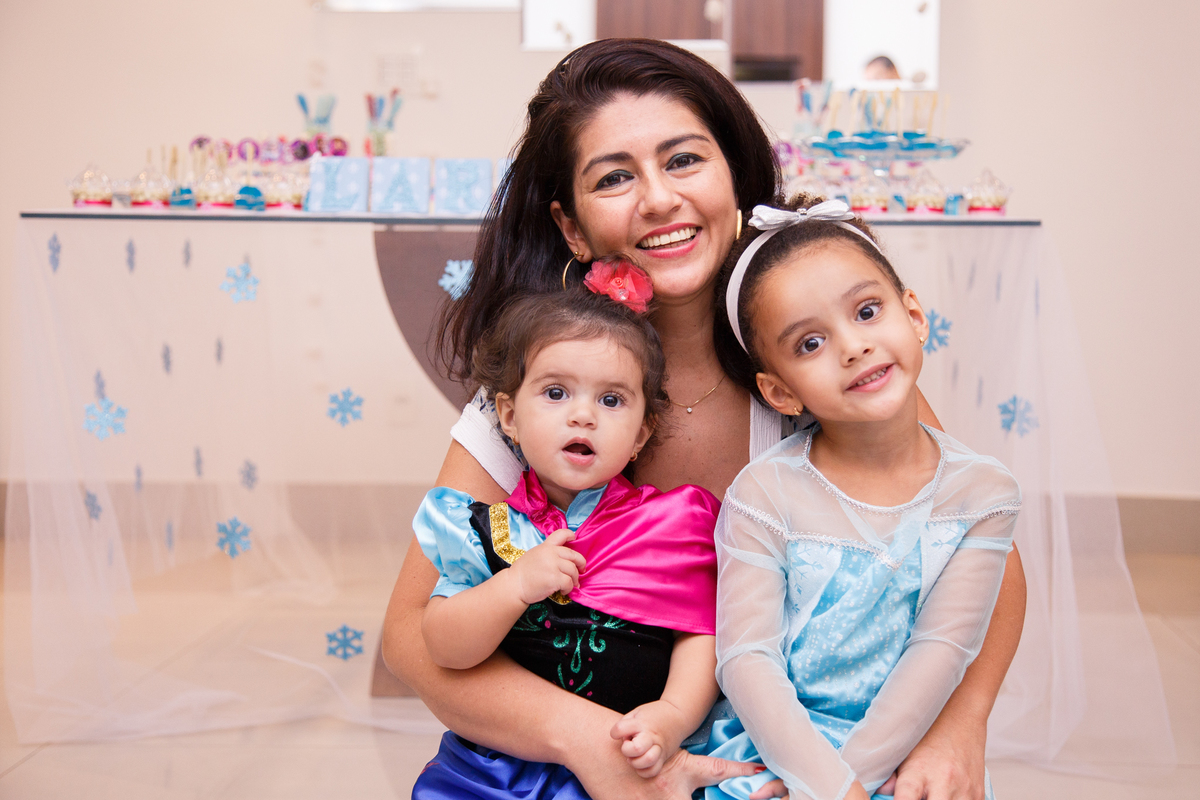 Crianças fantasiadas de Anna e Elsa do Frozen em frente à mesa do bolo do aniversário de 1 ano da mais nova, posando junto com sua mãe.