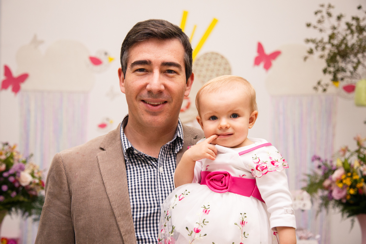 Pai com sua bebê aniversariante no colo, ambos olhando para a câmera, comemorando o aniversário da pequena.