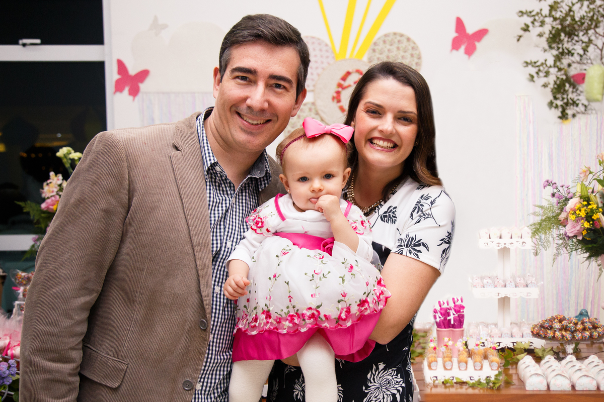 Pais com sua bebê no colo posando para foto em frente à mesa do bolo.