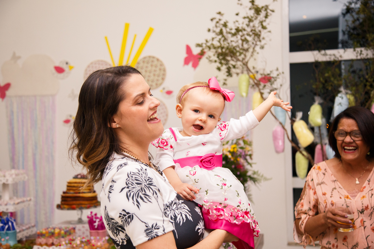Mãe com sua bebê no colo que está brincando, rindo com o braço para cima. A avó ri ao fundo da foto.