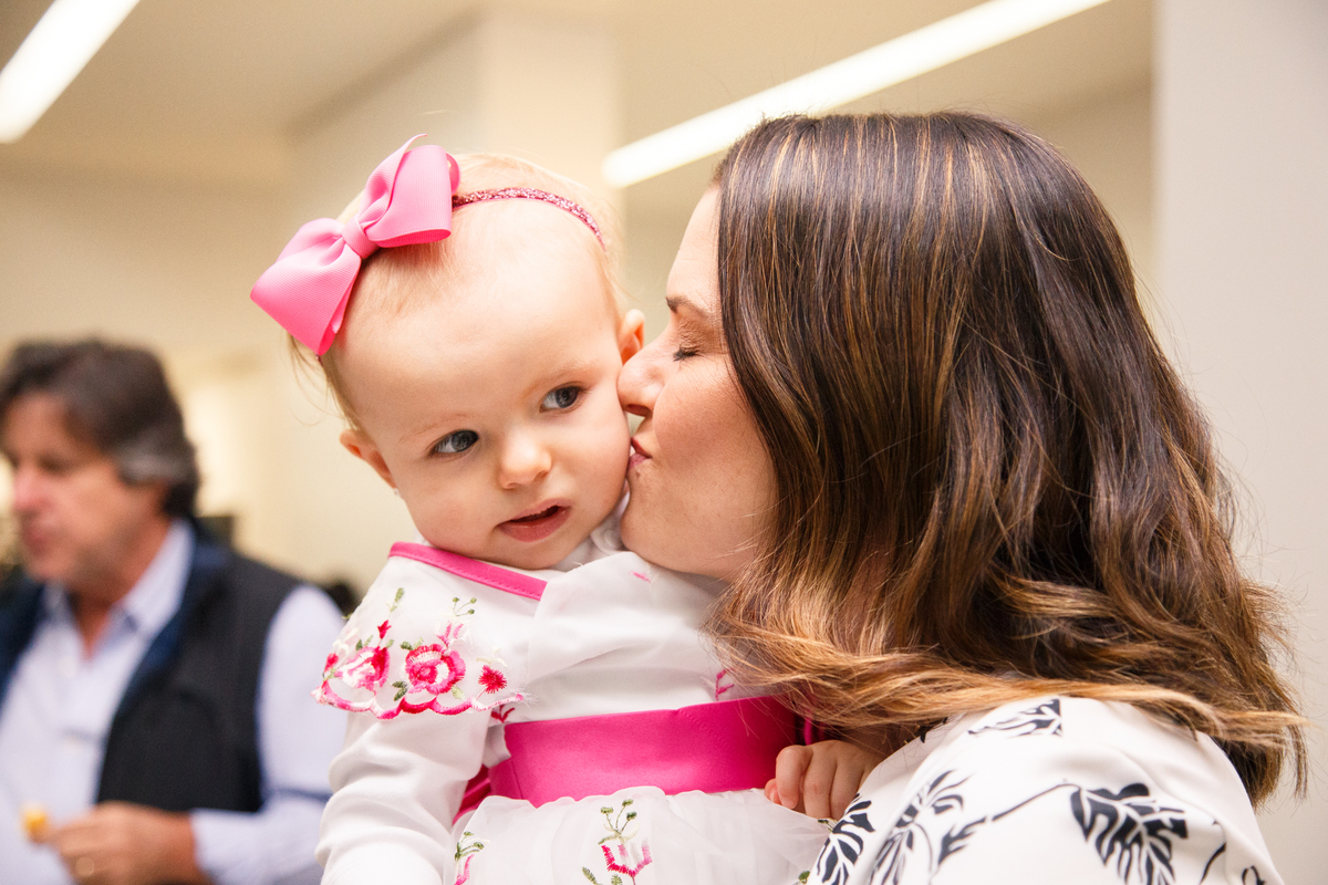 Mãe dá um beijo em sua bebê, que está em seu colo, durante festa de aniversário.